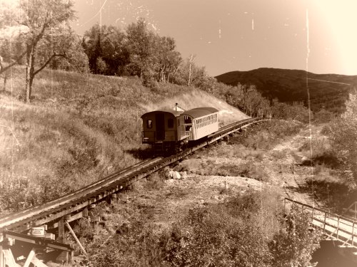 A vintage train travels along a curved track through a scenic landscape of trees and hills.