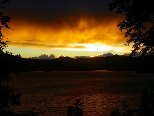 Sunset over a lake, with vibrant orange and yellow clouds reflecting on the water, framed by dark silhouettes of trees.