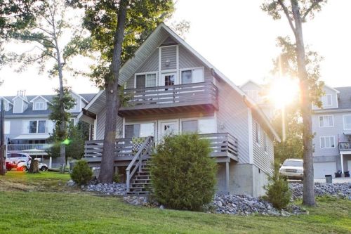 A cozy two-story house with a deck, surrounded by trees and grass, illuminated by the setting sun.