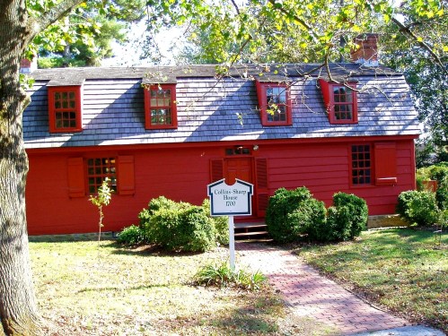 A red historic house with a shingled roof, surrounded by greenery and a sign reading "Collins Sharp House 1700."