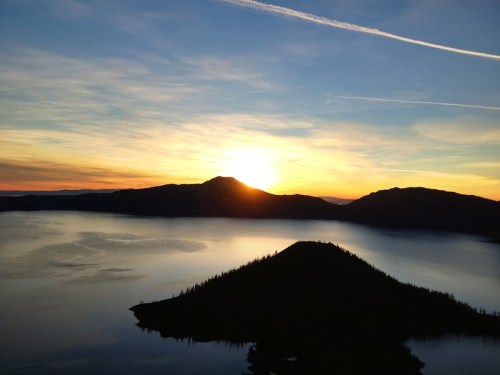 Sunset over a tranquil lake, with silhouettes of mountains and a small island in the foreground.
