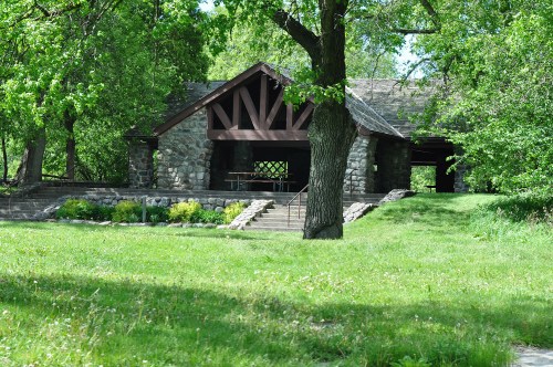 A stone pavilion surrounded by greenery, featuring a picnic table and steps leading up to it.