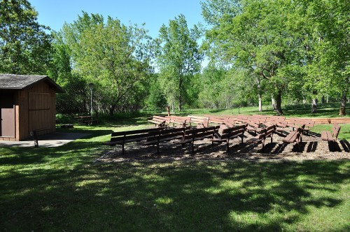 A grassy area with wooden benches arranged in rows, surrounded by trees and a small building in the background.
