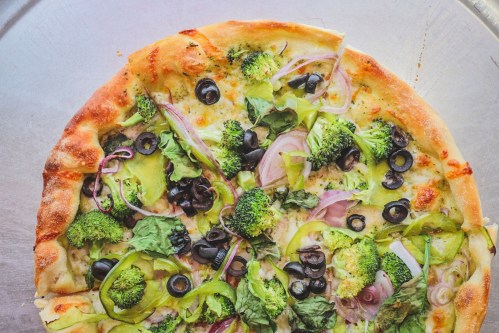 A freshly baked pizza topped with broccoli, olives, onions, and various greens on a silver tray.