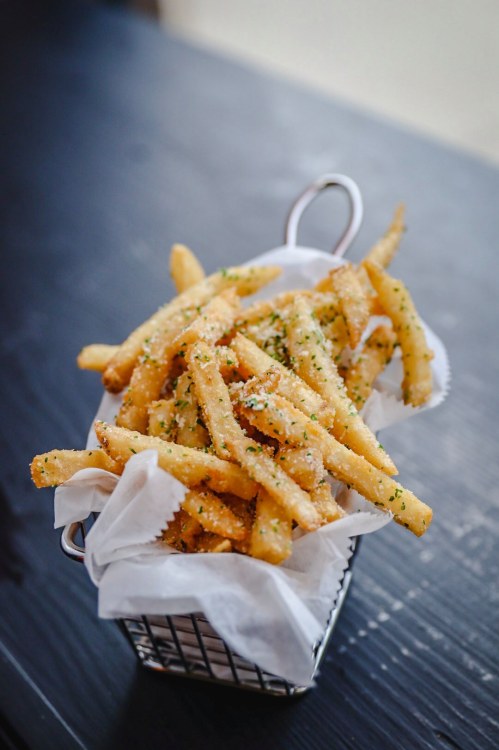 A basket of golden, crispy French fries sprinkled with herbs on a dark wooden surface.