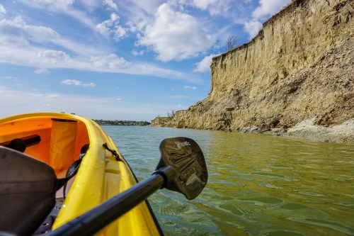 A yellow kayak on calm water, with a steep, rocky cliff and blue sky in the background.