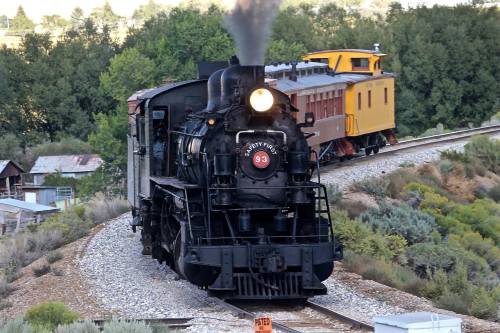A vintage steam locomotive with a black engine and yellow caboose, traveling along a curved track surrounded by greenery.