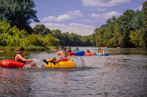 People enjoying a sunny day kayaking and tubing on a calm river surrounded by lush greenery.
