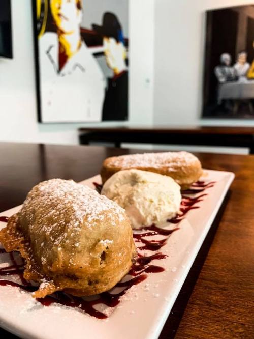Three fried desserts dusted with powdered sugar, served with a scoop of ice cream on a plate drizzled with sauce.