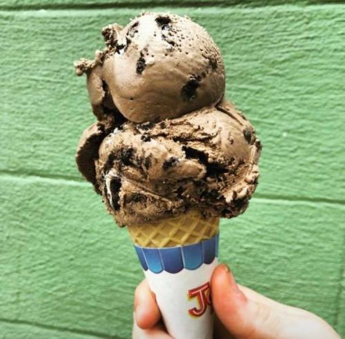 A hand holding a cone with two scoops of chocolate ice cream with cookie pieces, against a green wall.
