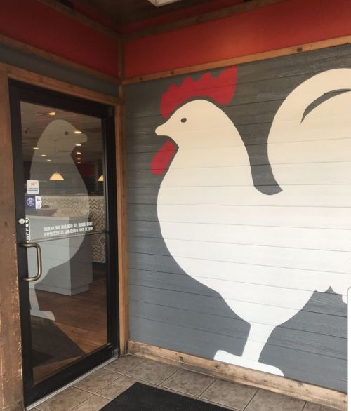 A large white rooster mural on a gray wall next to a glass door of a restaurant.