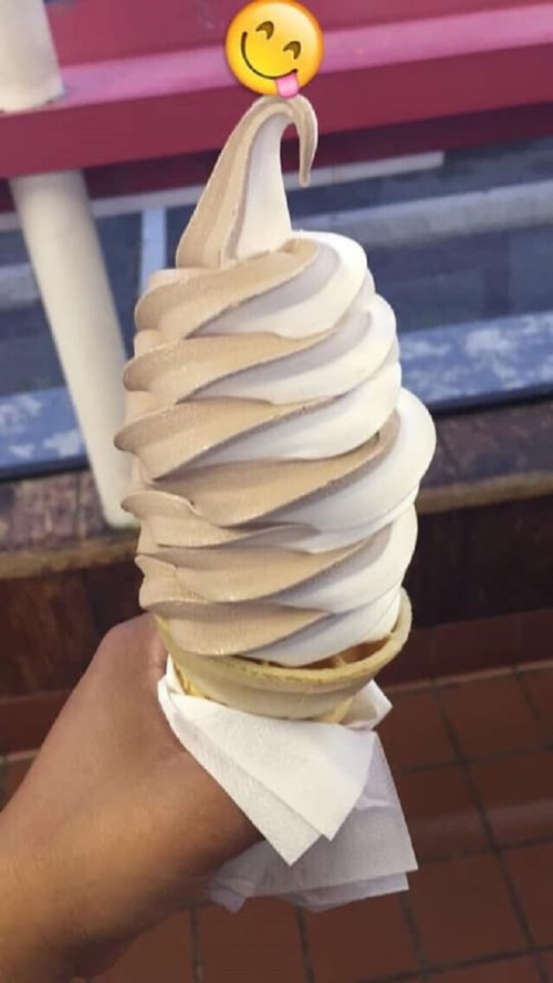 A hand holding a swirled soft serve ice cream cone with chocolate and vanilla flavors, topped with a playful emoji.