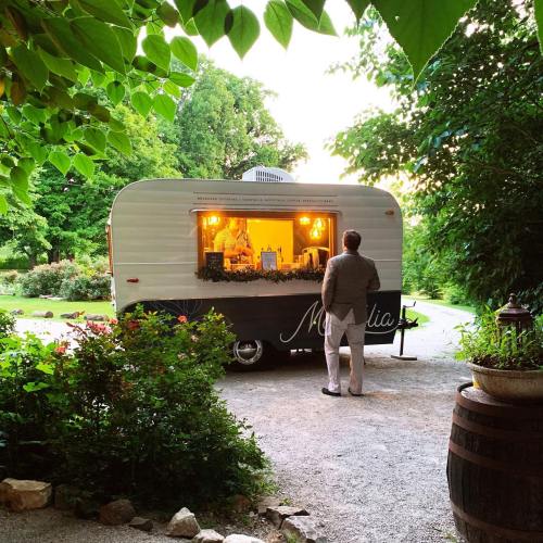 A person stands in front of a vintage trailer, illuminated by warm lights, surrounded by greenery and flowers.