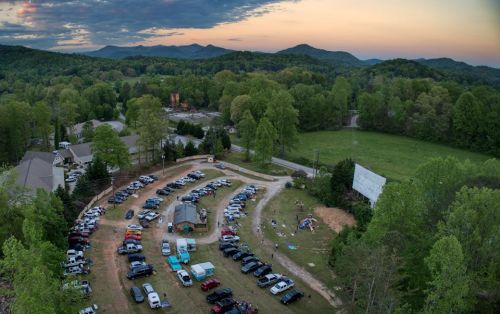 Aerial view of a parking area filled with cars, surrounded by trees and mountains at sunset.