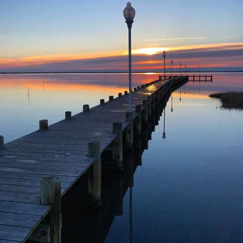 A wooden pier extends into calm waters at sunset, with lampposts lining the path and colorful reflections in the water.