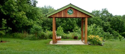 A wooden shelter in a park surrounded by lush greenery and yellow flowers, with a sign indicating the park's name.