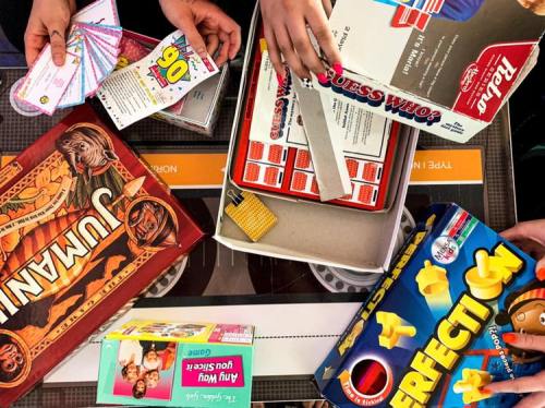 A tabletop scene featuring various board games, cards, and game pieces, with hands reaching for items.