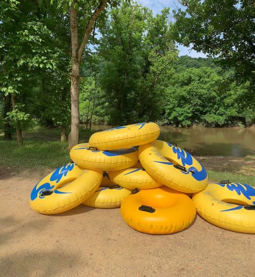 A stack of yellow and blue inflatable tubes beside a river, surrounded by green trees and a sandy area.