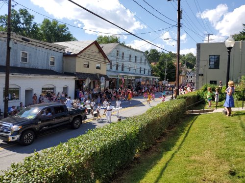 A lively parade on a sunny day, with crowds lining the street and a truck passing by in a small town.