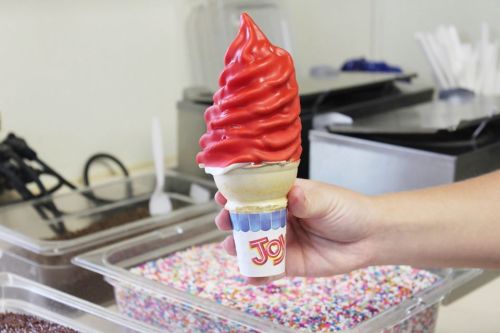 A hand holding a red soft-serve ice cream cone in front of a colorful sprinkle-filled container.