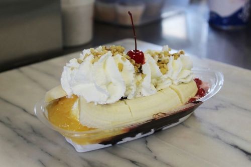 A banana split topped with whipped cream, nuts, cherries, and fruit sauces in a clear dish on a marble surface.