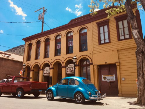 A bright blue vintage car parked in front of a yellow historic building with arched windows and a "Coffee Bar" sign.