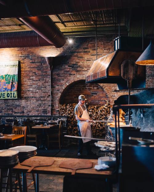 A chef in an apron stands in a rustic restaurant with exposed brick walls and a wood-fired oven.