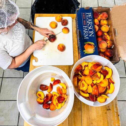 A person slices peaches on a cutting board, with bowls of sliced peaches and a box of whole peaches nearby.