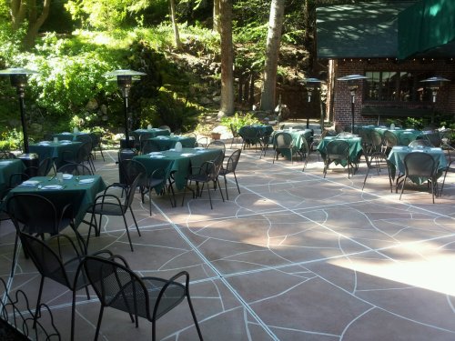 Outdoor dining area with green tables and chairs, surrounded by trees and a stone backdrop.
