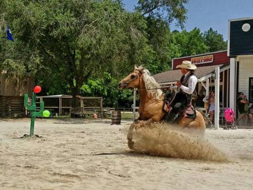 A cowboy in a hat rides a horse, kicking up sand while aiming at colorful targets in a western-themed setting.