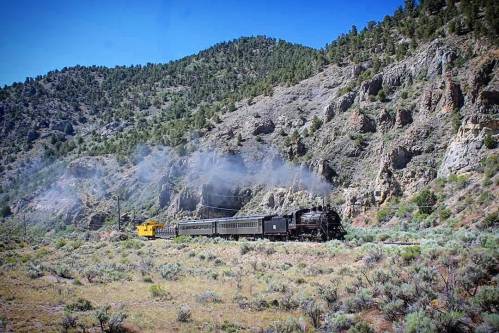 A vintage steam train travels through a mountainous landscape, with smoke billowing against a clear blue sky.