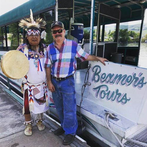 A man in traditional attire holds a drum next to a man in a plaid shirt by a boat labeled "Beamer's Tours."