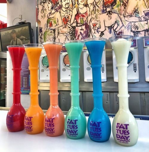 Colorful frozen drinks in tall glasses, lined up on a counter with a vibrant mural in the background.