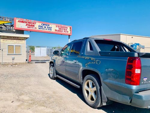 A gray SUV parked in front of an amusement park sign advertising drive-in movies on weekends. Clear blue sky above.