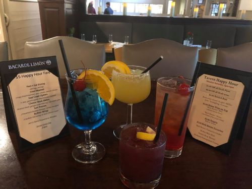 Five colorful cocktails on a table, with menus for happy hour specials in the background.