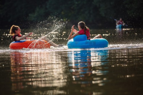 Two girls in colorful inner tubes splash water while floating on a calm river, enjoying a sunny day.