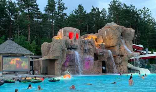 A pool with a rock formation and waterfall, featuring a movie screen and people swimming and relaxing in the water.