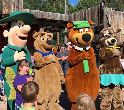 Four costumed bear characters stand with children at an outdoor event, smiling and engaging with the audience.