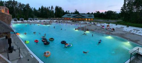 A vibrant pool scene at dusk, with people swimming and floating on colorful inflatables surrounded by lounge chairs.