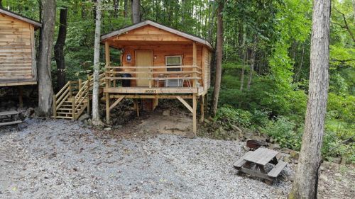 A wooden cabin on stilts surrounded by trees, with a picnic table and gravel area in front.