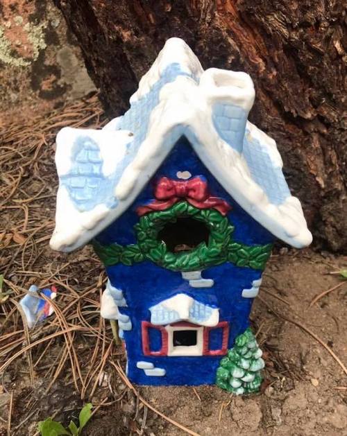 A small blue ceramic house with a snowy roof, decorated with a wreath and a tiny tree, nestled in the ground.
