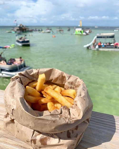 A paper bag filled with golden fries sits on a wooden railing, with boats and water in the background.