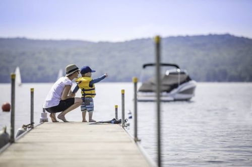 A woman and a child in a life jacket stand on a dock, pointing at a boat on the water with hills in the background.