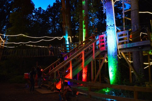 A brightly lit treehouse with colorful lights, surrounded by trees at dusk, with people enjoying the space.