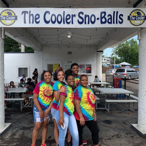 A group of five people in tie-dye shirts smiles in front of "The Cooler Sno-Balls" shop.