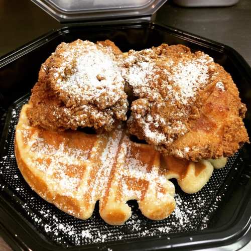 A close-up of a takeout container with two pieces of fried chicken on a waffle, dusted with powdered sugar.