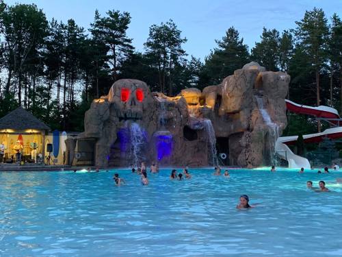 A lively pool scene with a rock formation featuring waterfalls and colorful lights, surrounded by trees and people swimming.