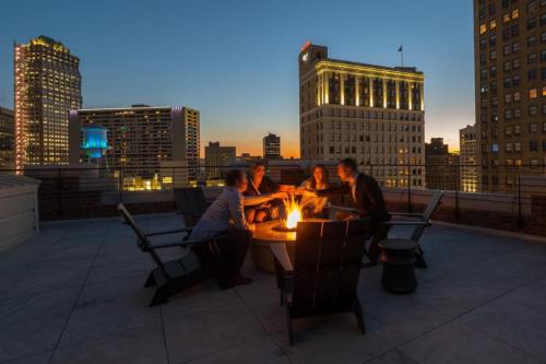A group of four people gathered around a fire pit on a rooftop at sunset, with city lights in the background.