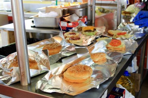 A food preparation station with several sesame seed burger buns and toppings wrapped in foil on a metal counter.