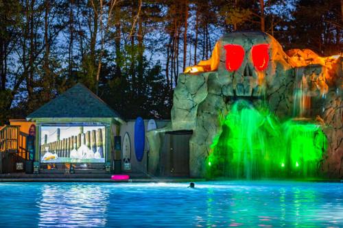 A pool area at night featuring a rock formation with a skull, colorful lights, and a large screen showing a movie.